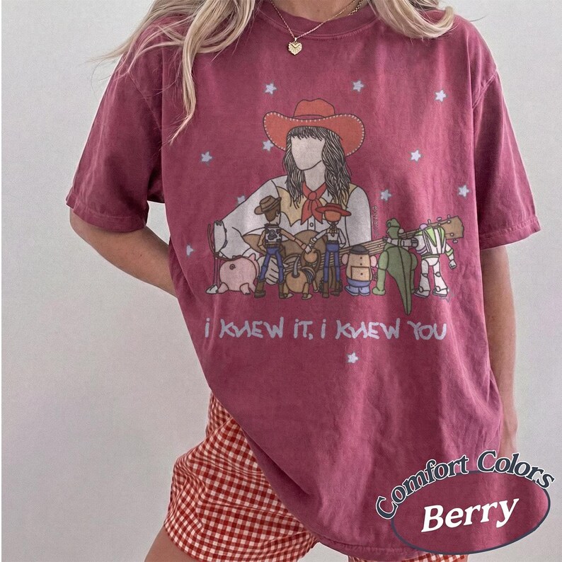 May include: A berry-coloured short-sleeve t-shirt featuring a graphic of a person in a cowboy hat playing a guitar surrounded by animated toy characters. The text "I KNEW IT, I KNEW YOU" is printed below the graphic, accented by small white stars.