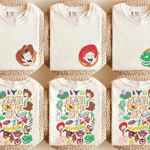 K&ouml;nnte beinhalten: Cremefarbene T-Shirts mit Toy Story Charaktergrafiken. Die obere Reihe zeigt die Gesichter von Woody, Jessie und Rex. Die untere Reihe zeigt eine Collage aus Charakteren und den Text "REACH FOR THE SKY" in einem farbenfrohen Design.