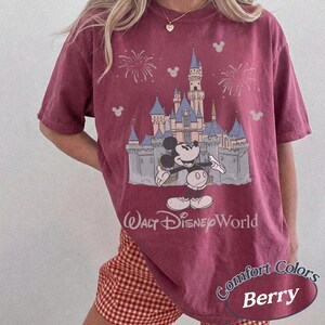 May include: Berry-colored t-shirt featuring a graphic of Mickey Mouse in front of a castle with fireworks and the words "Walt Disney World." The shirt also has the words "Comfort Colors" and "Berry" on it.
