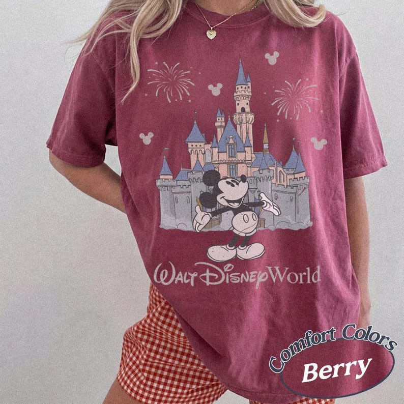 May include: Berry-colored t-shirt featuring a graphic of Mickey Mouse in front of a castle with fireworks and the words "Walt Disney World." The shirt also has the words "Comfort Colors" and "Berry" on it.