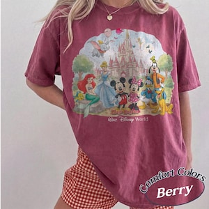 May include: A berry-colored short-sleeve t-shirt featuring a graphic print of various cartoon characters in front of a castle. The text "Magic Kingdom" and "Walt Disney World" is printed below the characters. The shirt has a crew neckline.