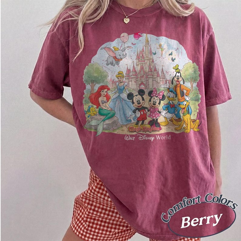 May include: A berry-colored short-sleeve t-shirt featuring a graphic print of various cartoon characters in front of a castle. The text "Magic Kingdom" and "Walt Disney World" is printed below the characters. The shirt has a crew neckline.