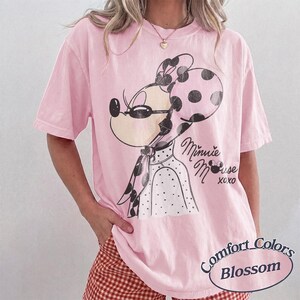 May include: A light pink Comfort Colors Blossom t-shirt featuring a Minnie Mouse graphic. The graphic includes Minnie Mouse wearing sunglasses and a polka dot bow and scarf. The text "Minnie Mouse xoxo" is also on the shirt.