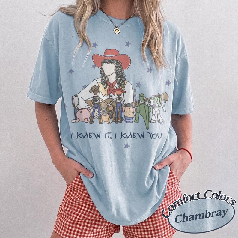 May include: A light blue t-shirt featuring a graphic illustration of a person in a red cowboy hat surrounded by various toy characters. The text "I KNEW IT, I KNEW YOU" is printed in dark blue below the illustration, accented by small purple stars.