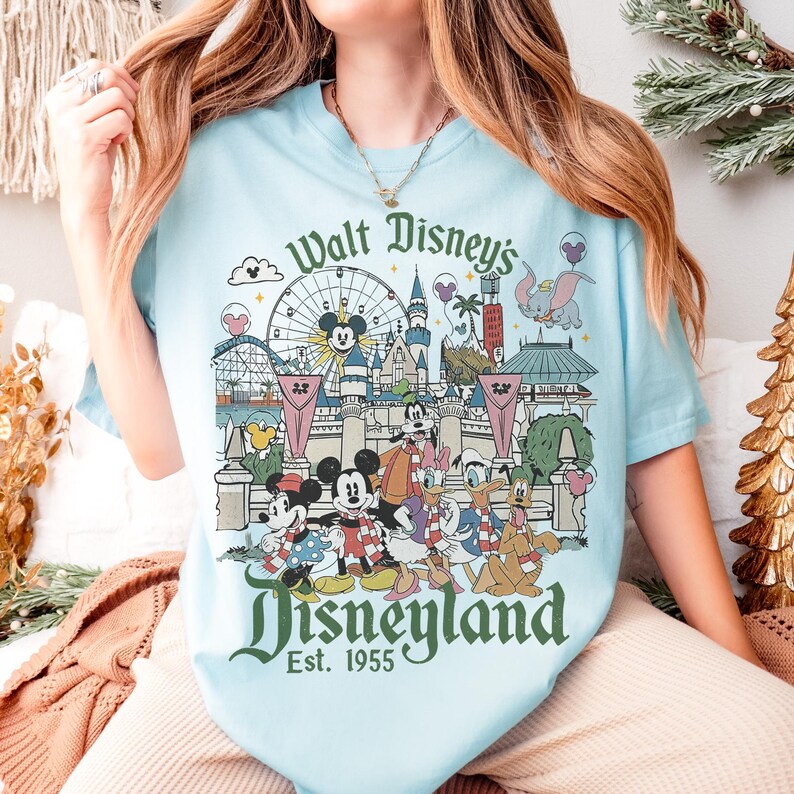 May include: Light blue t-shirt featuring a Disneyland graphic. The design includes the Disneyland logo, the phrase "Walt Disney's Disneyland Est. 1955", and various Disney characters and park attractions. The shirt is a soft, comfortable material.