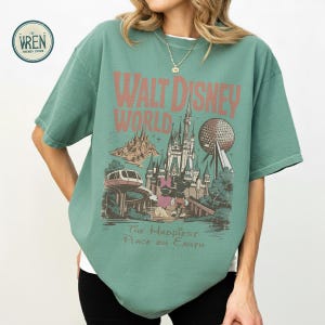 Może przedstawiać: Zielona koszulka z grafiką w stylu vintage z Walt Disney World. Projekt zawiera nazwę parku, zamek, Spaceship Earth i frazę "The Happiest Place on Earth."