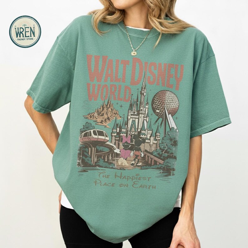 Może przedstawiać: Zielona koszulka z grafiką w stylu vintage z Walt Disney World. Projekt zawiera nazwę parku, zamek, Spaceship Earth i frazę "The Happiest Place on Earth."
