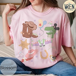 May include: Pink t-shirt with a colorful graphic featuring cartoon characters, including a cowboy hat, boots, a dinosaur, and a spaceship. The word "Blossom" is printed at the bottom of the shirt. The shirt is from the Wren Trendy Store.