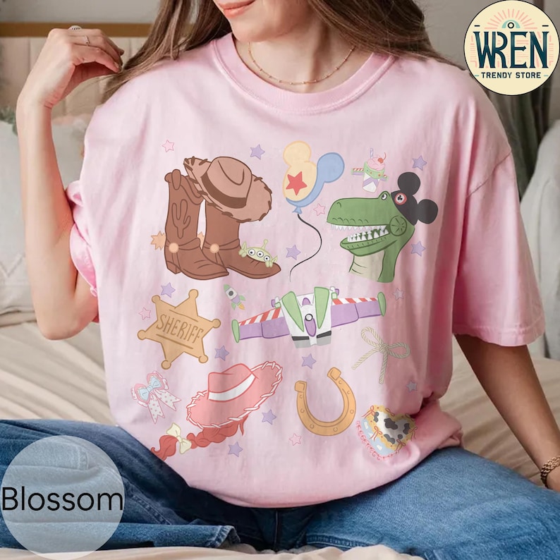 May include: Pink t-shirt with a colorful graphic featuring cartoon characters, including a cowboy hat, boots, a dinosaur, and a spaceship. The word "Blossom" is printed at the bottom of the shirt. The shirt is from the Wren Trendy Store.