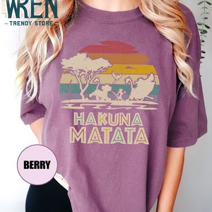 May include: Purple t-shirt with a vintage sunset graphic featuring silhouettes of characters from the Lion King movie. The text "Hakuna Matata" is printed below the graphic.