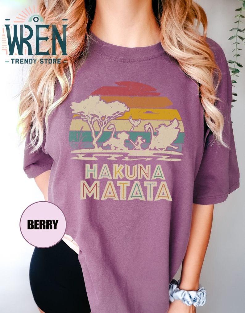 May include: Purple t-shirt with a vintage sunset graphic featuring silhouettes of characters from the Lion King movie. The text "Hakuna Matata" is printed below the graphic.