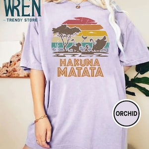 May include: A purple t-shirt with a vintage-style sunset graphic featuring a scene from the Lion King movie. The graphic includes a silhouette of Simba, Timon, and Pumbaa running through the savanna. The text "Hakuna Matata" is printed below the graphic.