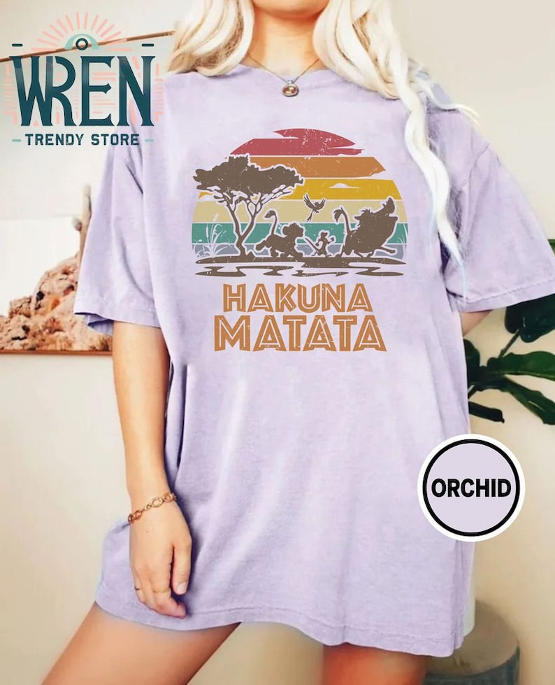 May include: A purple t-shirt with a vintage-style sunset graphic featuring a scene from the Lion King movie. The graphic includes a silhouette of Simba, Timon, and Pumbaa running through the savanna. The text "Hakuna Matata" is printed below the graphic.