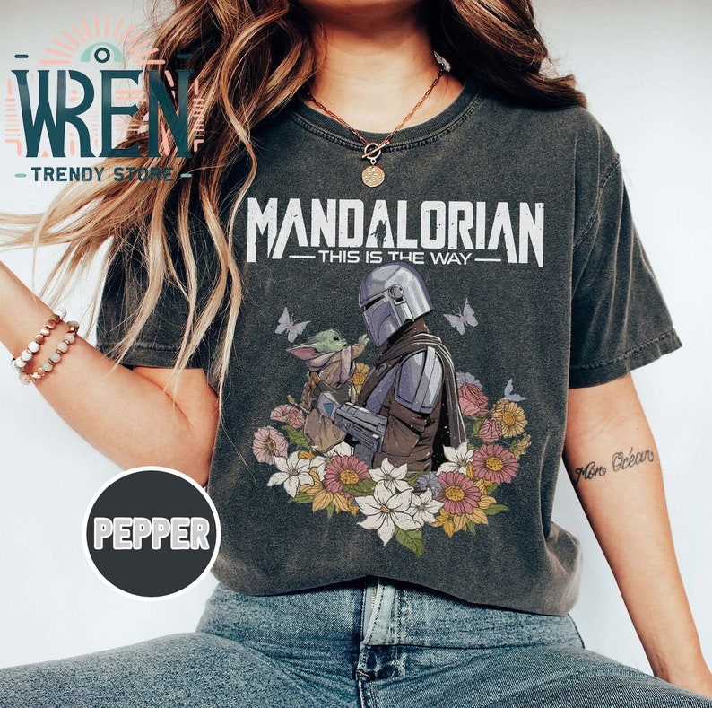 May include: A grey t-shirt with a distressed look featuring a graphic of the Mandalorian and Grogu from the Star Wars series. The graphic includes the text "Mandalorian - This is the Way -" and a floral design.