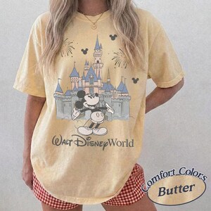 May include: Butter yellow t-shirt featuring a graphic of Mickey Mouse in front of Cinderella's castle at Walt Disney World. The shirt has a vintage look with the words "Walt Disney World" printed below the graphic. The shirt is a Comfort Colors brand.