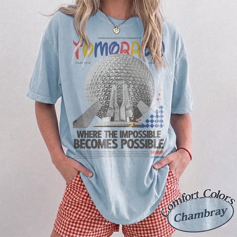 May include: A light blue short-sleeve t-shirt featuring a graphic of the Epcot Spaceship Earth structure. The text reads TOMORROW, SPARK YOUR, WHERE THE IMPOSSIBLE BECOMES POSSIBLE, and EXPERIMENTAL PROTOTYPE COMMUNITY OF TOMORROW in various colors.
