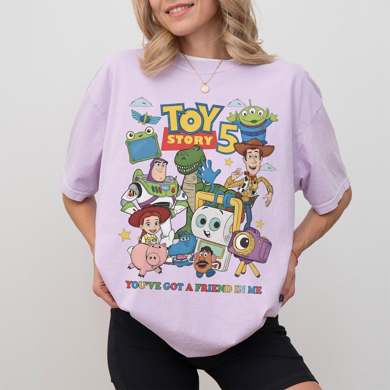 May include: A light purple t-shirt featuring a colorful graphic of characters from the animated movie "Toy Story 5". The design includes Woody, Buzz Lightyear, and other characters, with the text "YOU'VE GOT A FRIEND IN ME".