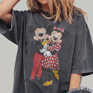 May include: A charcoal gray short-sleeve t-shirt featuring a distressed graphic of Mickey and Minnie Mouse embracing. Mickey wears a tuxedo with a yellow bowtie, and Minnie wears a red polka-dot dress with yellow shoes. Text reads Comfort Colors Pepper.