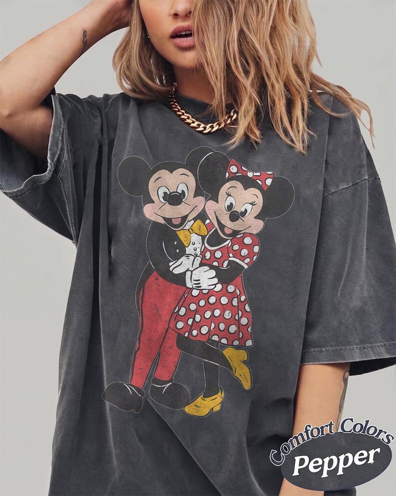 May include: A charcoal gray short-sleeve t-shirt featuring a distressed graphic of Mickey and Minnie Mouse embracing. Mickey wears a tuxedo with a yellow bowtie, and Minnie wears a red polka-dot dress with yellow shoes. Text reads Comfort Colors Pepper.