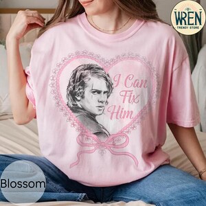 May include: Pale pink t-shirt featuring a portrait of a man within a heart-shaped frame, accompanied by the text "I Can Fix Him". The design incorporates a pink bow and lace detailing. Part of the "Blossom" collection.