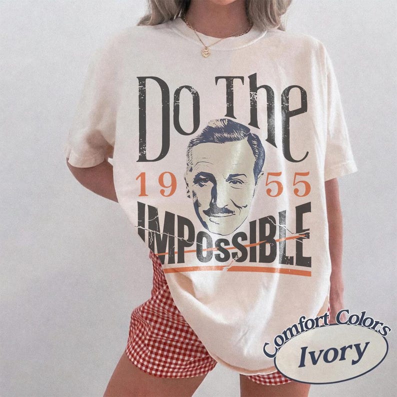 May include: Ivory-colored t-shirt with the text "Do The Impossible" and the numbers "1955". The shirt features a vintage image of a man's face. The shirt is labeled "Comfort Colors Ivory".