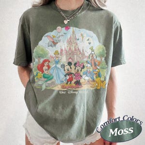 May include: A moss green t-shirt featuring a graphic print of various cartoon characters in front of a castle. The text "Magic Kingdom" and "Walt Disney World" is printed below the characters. The shirt has short sleeves and a crew neckline.