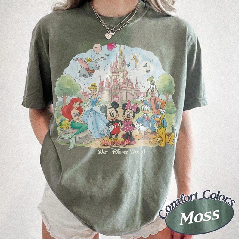 May include: A moss green t-shirt featuring a graphic print of various cartoon characters in front of a castle. The text "Magic Kingdom" and "Walt Disney World" is printed below the characters. The shirt has short sleeves and a crew neckline.
