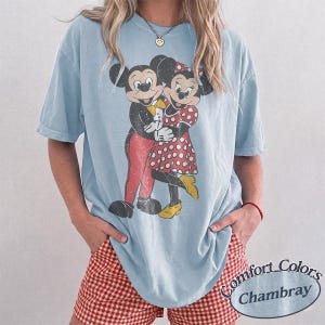 May include: A light blue short-sleeve t-shirt featuring a distressed graphic print of Mickey and Minnie Mouse embracing. The shirt has a crew neckline and a relaxed fit. The graphic shows Mickey in red pants and Minnie in a red polka dot dress.