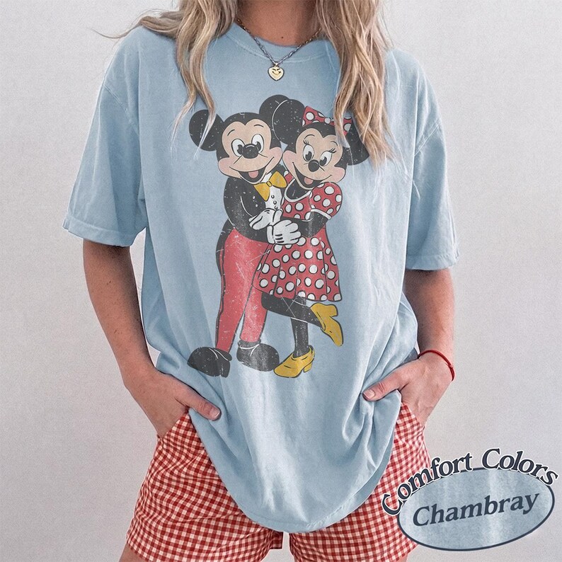 May include: A light blue short-sleeve t-shirt featuring a distressed graphic print of Mickey and Minnie Mouse embracing. The shirt has a crew neckline and a relaxed fit. The graphic shows Mickey in red pants and Minnie in a red polka dot dress.