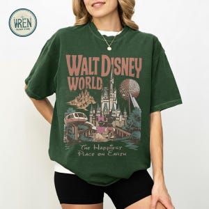 Może przedstawiać: Ciemnozielona koszulka z grafiką w stylu vintage z Walt Disney World. Projekt zawiera nazwę parku, zamek, Spaceship Earth i frazę "The Happiest Place on Earth."