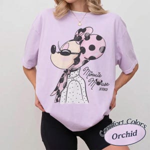 May include: A light purple t-shirt featuring a Minnie Mouse graphic with a pink polka dot bow and scarf. The text "Minnie Mouse xoxo" is below the graphic. The shirt is labeled "Comfort Colors Orchid".