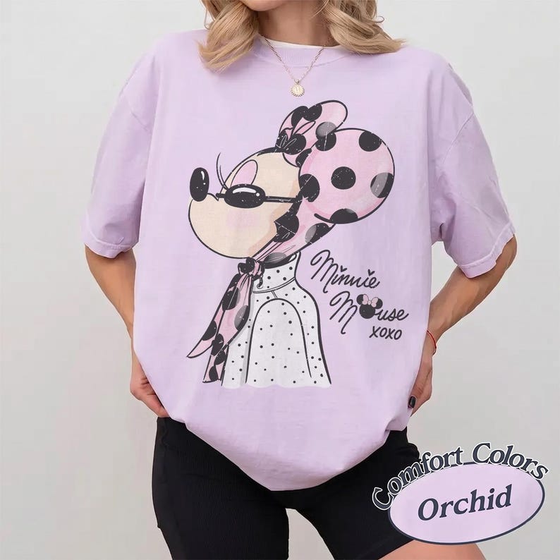 May include: A light purple t-shirt featuring a Minnie Mouse graphic with a pink polka dot bow and scarf. The text "Minnie Mouse xoxo" is below the graphic. The shirt is labeled "Comfort Colors Orchid".