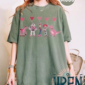 Może przedstawiać: Zielony T-shirt z grafiką postaci z Toy Story: Woody, Buzz Astral, Jessie i Rex trzymających r&oacute;żowe balony w kształcie serca. Tekst "MOSS" znajduje się w prawym g&oacute;rnym rogu obrazu.