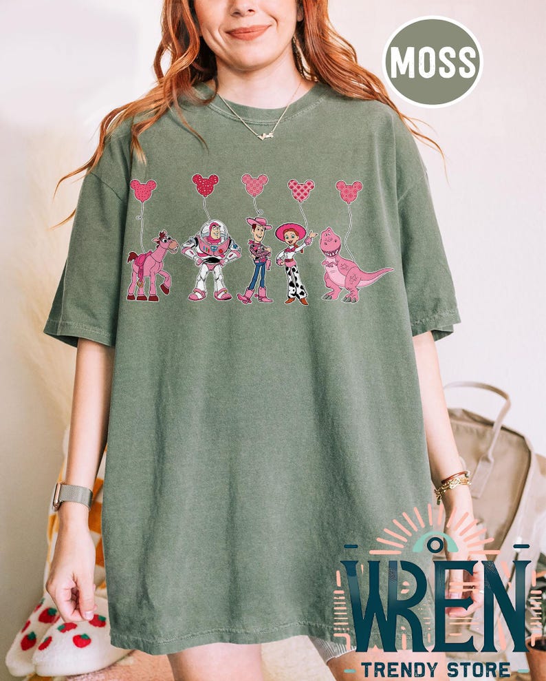 Może przedstawiać: Zielony T-shirt z grafiką postaci z Toy Story: Woody, Buzz Astral, Jessie i Rex trzymających r&oacute;żowe balony w kształcie serca. Tekst "MOSS" znajduje się w prawym g&oacute;rnym rogu obrazu.