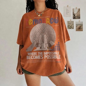 May include: An oversized burnt orange t-shirt featuring a large graphic of a geodesic dome structure. The text reads "TOMORROW" in colorful letters and "WHERE THE IMPOSSIBLE BECOMES POSSIBLE" in white, along with smaller printed details.