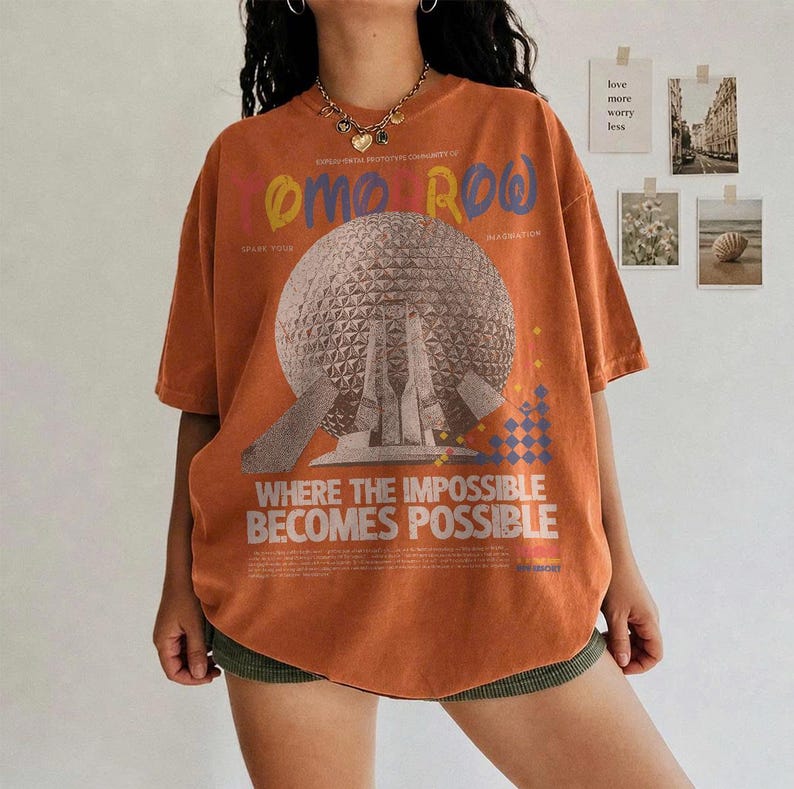 May include: An oversized burnt orange t-shirt featuring a large graphic of a geodesic dome structure. The text reads "TOMORROW" in colorful letters and "WHERE THE IMPOSSIBLE BECOMES POSSIBLE" in white, along with smaller printed details.