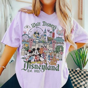 May include: A light purple t-shirt featuring a colorful Disneyland graphic. The graphic includes Mickey and Minnie Mouse, Donald Duck, and other Disney characters in front of Disneyland landmarks. Text reads "Walt Disney's Disneyland Est. 1955."