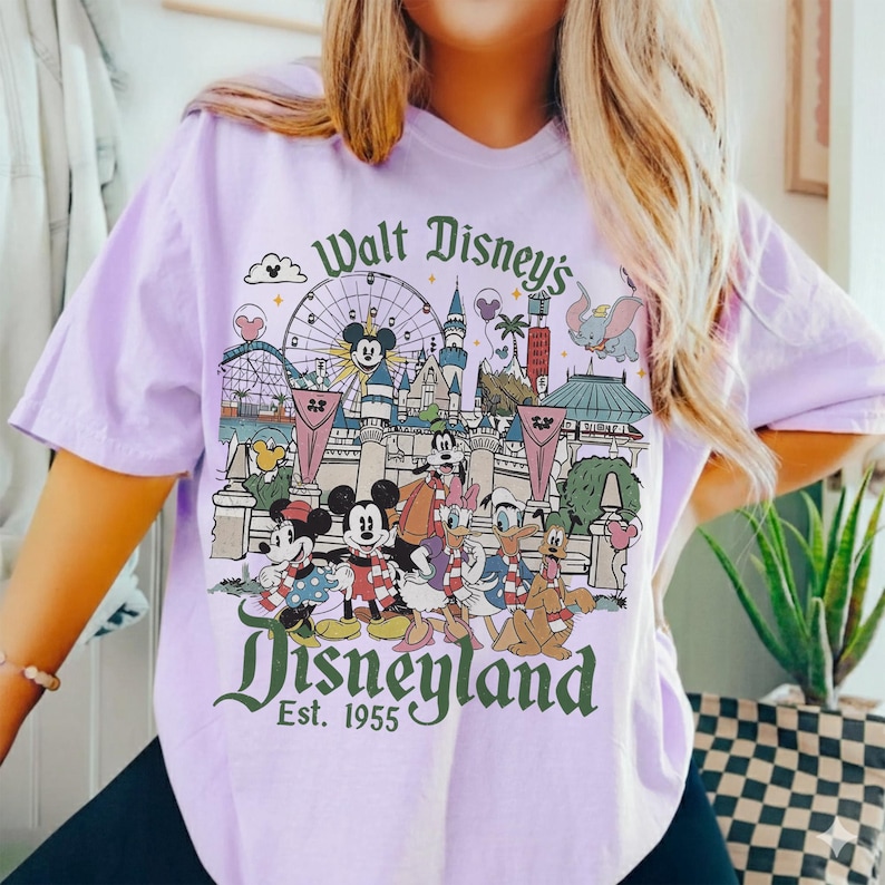 May include: A light purple t-shirt featuring a colorful Disneyland graphic. The graphic includes Mickey and Minnie Mouse, Donald Duck, and other Disney characters in front of Disneyland landmarks. Text reads "Walt Disney's Disneyland Est. 1955."