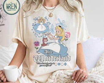 May include: Cream-colored t-shirt featuring a graphic of Alice in Wonderland. The design includes Alice, the Cheshire Cat, and floral elements. Text on the shirt reads "Alice in Wonderland" and "What kind of garden do you come from?"