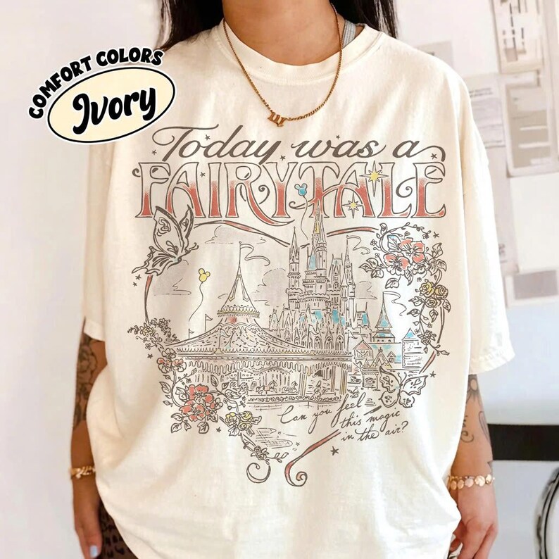 Puede incluir: Una camiseta de manga corta color marfil con un estampado gr&aacute;fico de un castillo, una mariposa y elementos florales. El texto dice Today was a FAIRY TALE y Can you feel this magic in the air? La etiqueta Comfort Colors Ivory es visible en la esquina.