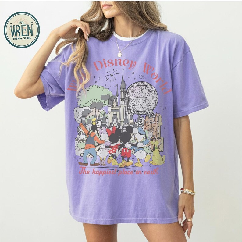以下が含まれることがあります： ディズニーワールドのグラフィックが特徴のライラック色のTシャツ。デザインには、ピンク色の「ディズニーワールド」の文字と、ディズニーのキャラクターや公園のランドマークのイラストが含まれています。「The happiest place on earth」というフレーズもプリントされています。
