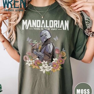 May include: Green t-shirt featuring a floral design and a graphic of the Mandalorian and Grogu. The text "Mandalorian - This is the Way -" is printed on the shirt.