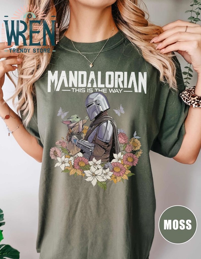 May include: Green t-shirt featuring a floral design and a graphic of the Mandalorian and Grogu. The text "Mandalorian - This is the Way -" is printed on the shirt.