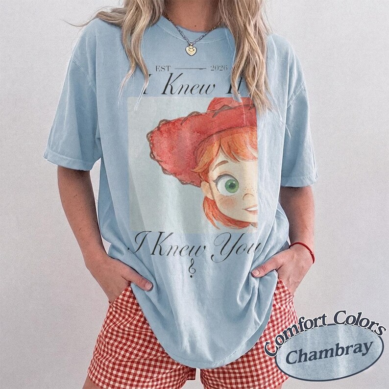 May include: A light blue short-sleeve t-shirt featuring a graphic of a red-haired character in a red cowboy hat. The text reads EST 2026 I Knew I Knew You. The shirt is paired with red and white gingham shorts. The fabric has a washed-out appearance.
