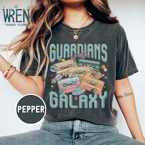 K&ouml;nnte beinhalten: Ein dunkelgraues T-Shirt mit einem Vintage-Grafikdesign, das den Text "Guardians of the Galaxy" und Kassetten mit Texten aus beliebten Songs zeigt. Das Design enth&auml;lt Sterne, Planeten und ein Raumschiff.