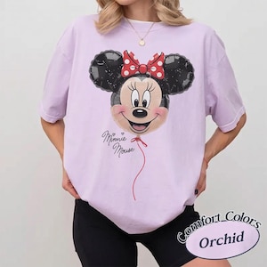 Op de afbeelding: Een lichtpaars T-shirt met een Minnie Mouse ballonontwerp, een rode strik en de tekst "Minnie Mouse". Het shirt is gelabeld "Comfort Colors Orchid".
