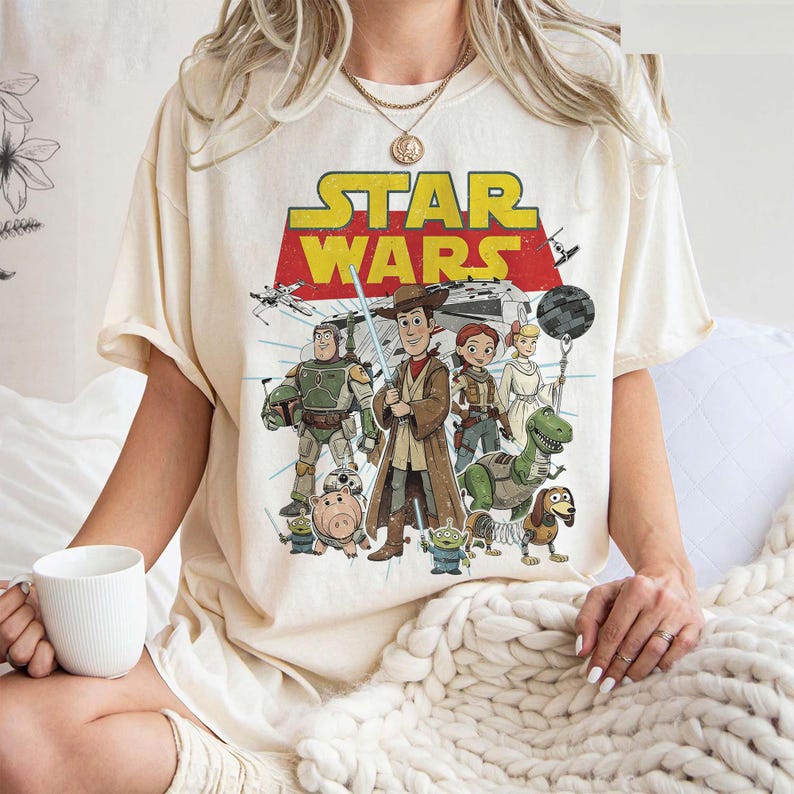 May include: Cream-colored t-shirt featuring a graphic of Star Wars characters. The design includes the Star Wars logo in yellow and red, with illustrations of various characters from the franchise. The shirt has short sleeves and a relaxed fit.