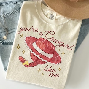 May include: A cream-colored t-shirt featuring a graphic of a red cowgirl hat and a red braided ponytail. The text "you're a Cowgirl like me" is printed in a dark red script font, surrounded by small gold star icons. The shirt has a Comfort Colors label.