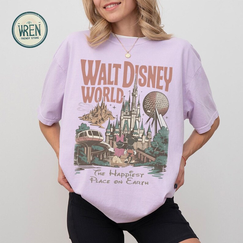 Może przedstawiać: Jasnofioletowa koszulka z grafiką Walt Disney World. Projekt zawiera tekst "Walt Disney World" i "The Happiest Place on Earth", a także ilustracje zamku, Spaceship Earth i innych atrakcji parku.