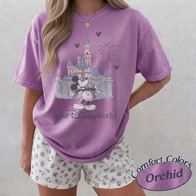 May include: Orchid-colored t-shirt featuring a graphic of Mickey Mouse in front of a castle with fireworks. The text "Walt Disney World" is below the graphic. The shirt is paired with white shorts with a blue floral pattern. The words "Comfort Colors Orchid" are in a purple oval.